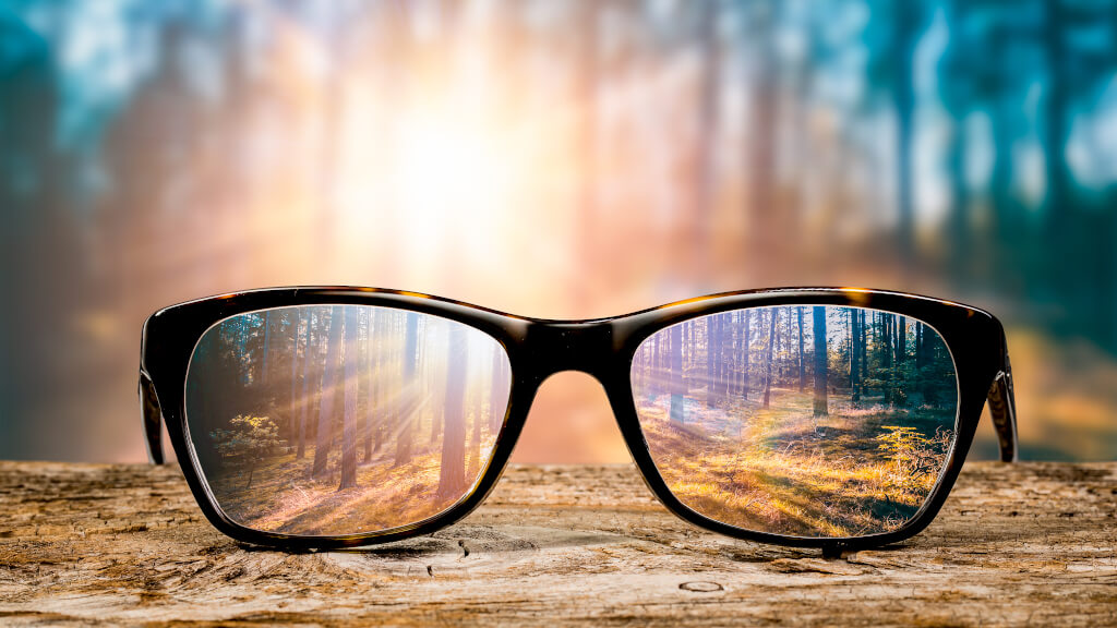Färgglad bild på et par glasögon som ligger på marken framför en skog. Allt är suddigt utom det man ser genom glasögonen som gör allt skarpt och fint.